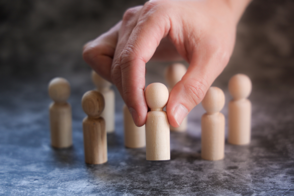 The hand of a Caucasian individual moving and rearranging wooden figurines that resemble people-shaped game pieces.
