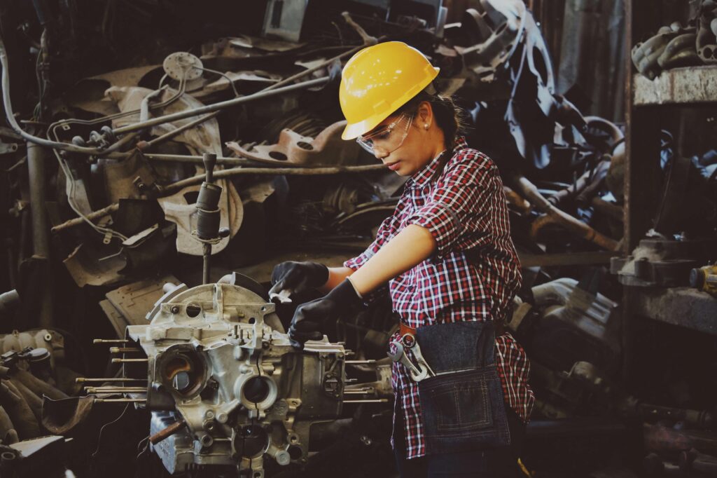 Woman with dark, braided hair wearing a yellow construction hat, plaid button-up, and tool belt working on building or repairing a machine part